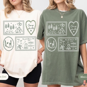 May include: Ivory and moss green t-shirts with white graphic designs. The designs include stamps with mountain, tree, and tent illustrations, along with text that reads "Last Trail Before the Veil" and "Camp Mille".