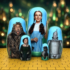 May include: A set of five light blue nesting dolls featuring painted portraits of characters from The Wizard of Oz, including Dorothy, the Cowardly Lion, the Scarecrow, the Tin Man, and Toto, arranged on a yellow brick pattern surface.