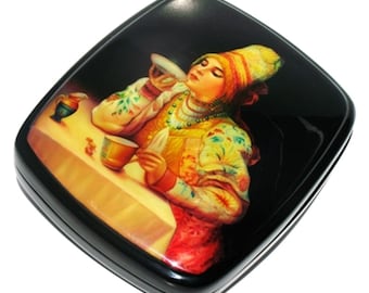 May include: A black lacquered wooden box with a painted scene of a woman in a colorful traditional Russian dress holding a cup and saucer. The woman is sitting at a table with a pitcher and other items on it.