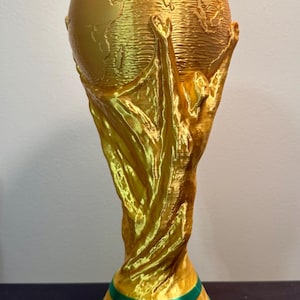 May include: A gold-coloured trophy featuring a globe supported by two figures with raised arms. The base is green with a gold band and the embossed text FIFA WORLD CUP. The structure has a visible layered texture from the 3D printing process.