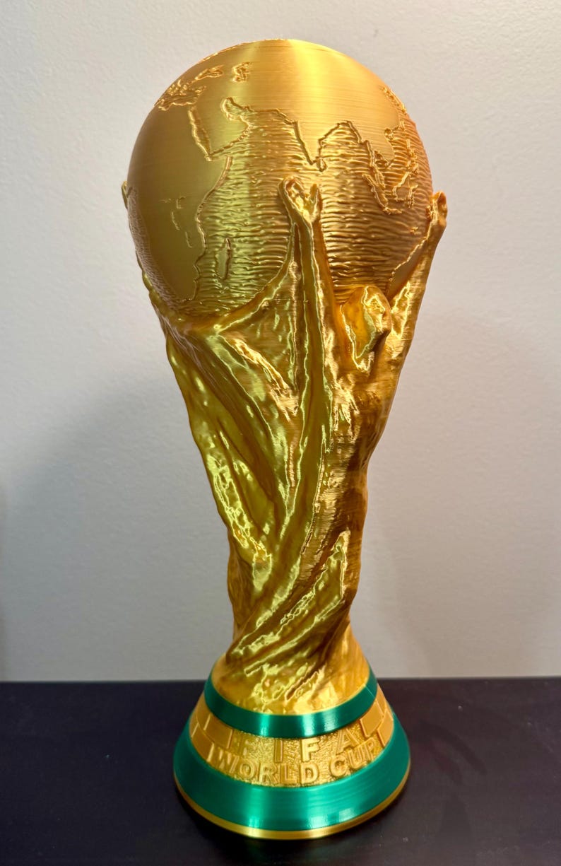 May include: A gold-coloured trophy featuring a globe supported by two figures with raised arms. The base is green with a gold band and the embossed text FIFA WORLD CUP. The structure has a visible layered texture from the 3D printing process.