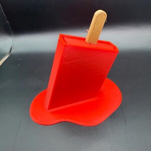 Peut inclure: Un support de t&eacute;l&eacute;phone rouge en forme de glace &agrave; l'eau avec un b&acirc;ton en bois. Le support est con&ccedil;u pour ressembler &agrave; une glace &agrave; l'eau fondante, avec la base rouge ressemblant &agrave; la friandise fondue. Le b&acirc;ton en bois est ins&eacute;r&eacute; dans le haut du support.