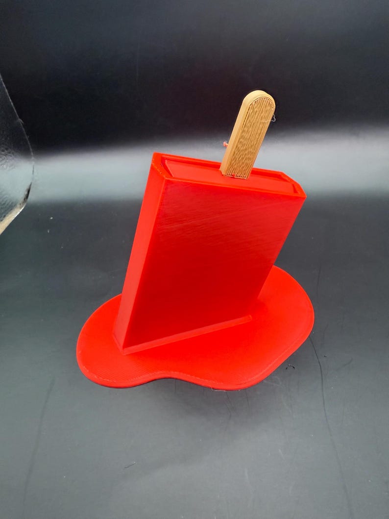 Peut inclure: Un support de t&eacute;l&eacute;phone rouge en forme de glace &agrave; l'eau avec un b&acirc;ton en bois. Le support est con&ccedil;u pour ressembler &agrave; une glace &agrave; l'eau fondante, avec la base rouge ressemblant &agrave; la friandise fondue. Le b&acirc;ton en bois est ins&eacute;r&eacute; dans le haut du support.