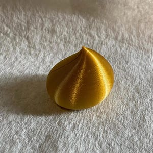 Könnte beinhalten: Ein goldfarbenes, tropfenförmiges Objekt mit strukturierter Oberfläche. Das Objekt hat eine spitze Spitze und eine abgerundete Basis. Die Oberfläche weist eine Reihe paralleler Linien auf, die von der Basis zur Spitze verlaufen. Das Objekt liegt auf einer weißen, strukturierten Oberfläche.