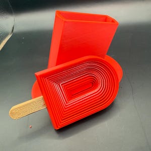 Peut inclure: Un objet en forme de glace, imprim&eacute; en 3D, de couleur rouge vif, avec un b&acirc;ton en bois. La glace a un design en couches, et un bloc rectangulaire est derri&egrave;re elle. Les objets sont sur une surface sombre.