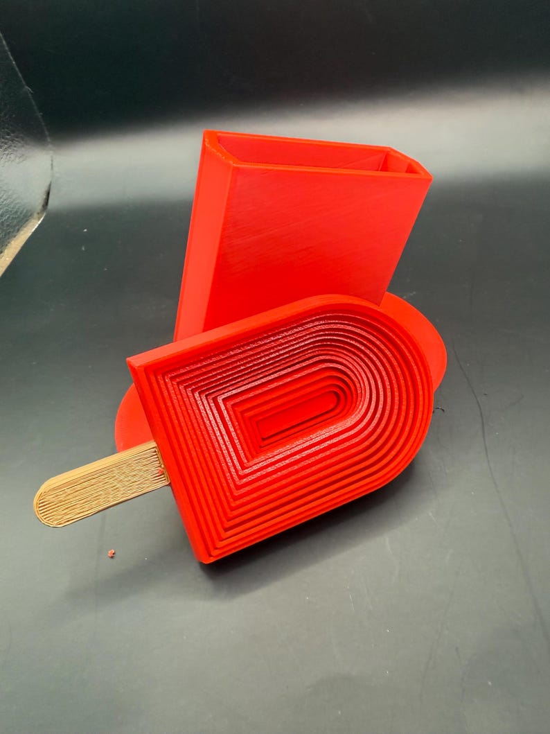 Peut inclure: Un objet en forme de glace, imprim&eacute; en 3D, de couleur rouge vif, avec un b&acirc;ton en bois. La glace a un design en couches, et un bloc rectangulaire est derri&egrave;re elle. Les objets sont sur une surface sombre.