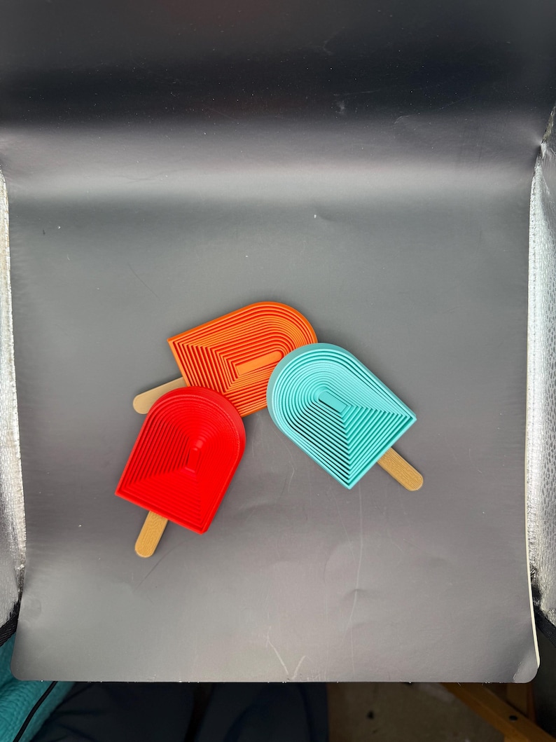 Peut inclure: Trois objets en forme de sucettes glac&eacute;es de couleurs vives : rouge, orange et turquoise. Chacun a un b&acirc;tonnet en bois et un design en couches. Les sucettes sont dispos&eacute;es sur une surface grise.