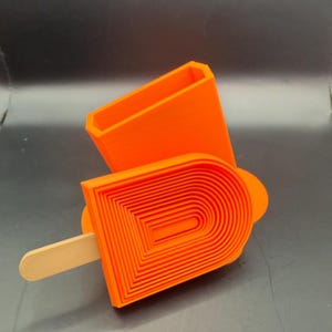 Peut inclure: Moule &agrave; popsicle orange vif avec un r&eacute;cipient rectangulaire assorti. Le moule &agrave; popsicle pr&eacute;sente un design en couches et un b&acirc;tonnet en bois. Le r&eacute;cipient est ouvert en haut. Les articles sont sur une surface gris fonc&eacute;.