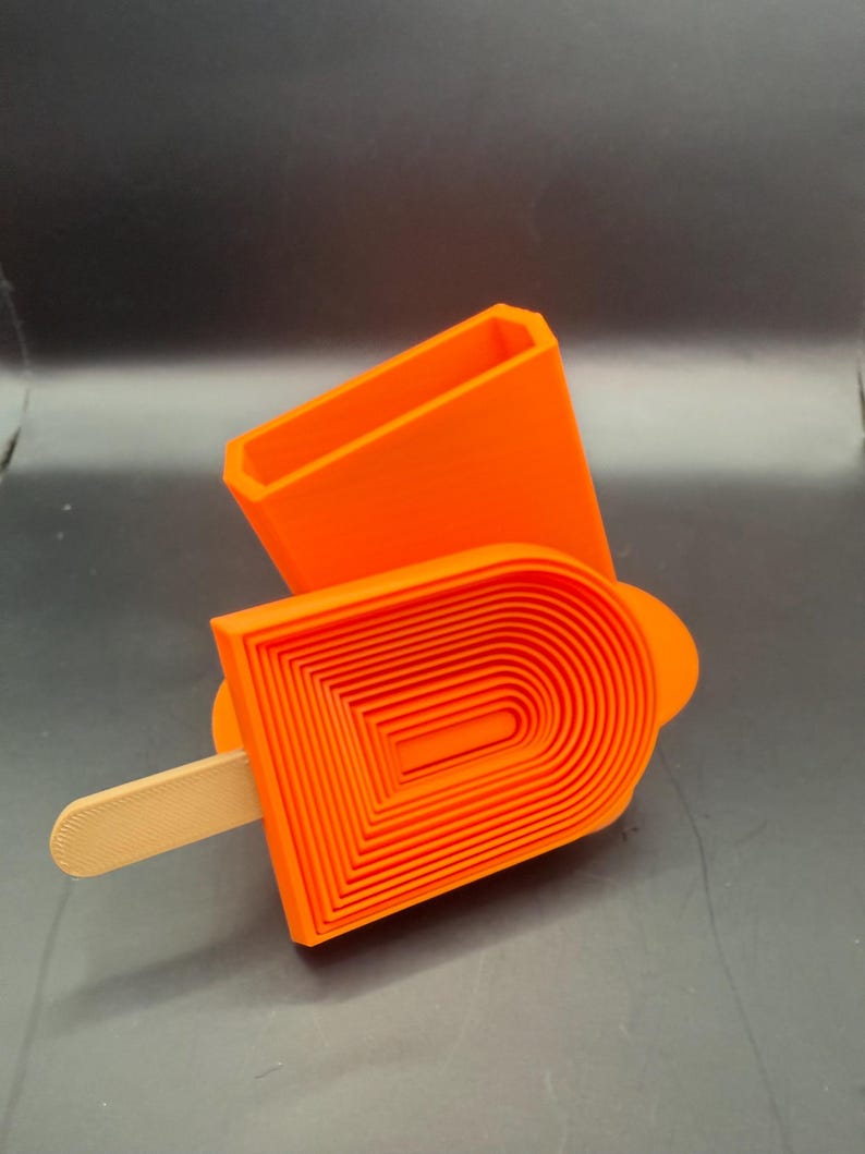 Peut inclure: Moule &agrave; popsicle orange vif avec un r&eacute;cipient rectangulaire assorti. Le moule &agrave; popsicle pr&eacute;sente un design en couches et un b&acirc;tonnet en bois. Le r&eacute;cipient est ouvert en haut. Les articles sont sur une surface gris fonc&eacute;.