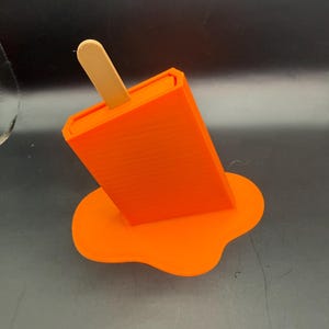 Peut inclure: Objet orange en forme de glace &agrave; l'eau avec un b&acirc;ton en bois, semblant fondre. La base de la glace est en forme de flaque, &eacute;galement orange. Il s'agit d'un objet imprim&eacute; en 3D.