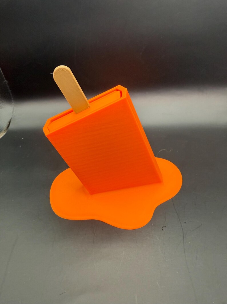 Peut inclure: Objet orange en forme de glace &agrave; l'eau avec un b&acirc;ton en bois, semblant fondre. La base de la glace est en forme de flaque, &eacute;galement orange. Il s'agit d'un objet imprim&eacute; en 3D.