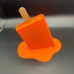 Peut inclure: Un support de t&eacute;l&eacute;phone en forme de glace &agrave; l'eau orange avec un b&acirc;ton en bois. Le support est con&ccedil;u pour ressembler &agrave; une glace &agrave; l'eau en train de fondre, avec la &laquo; cr&egrave;me glac&eacute;e &raquo; orange qui d&eacute;gouline sur la surface. Le support est en plastique.
