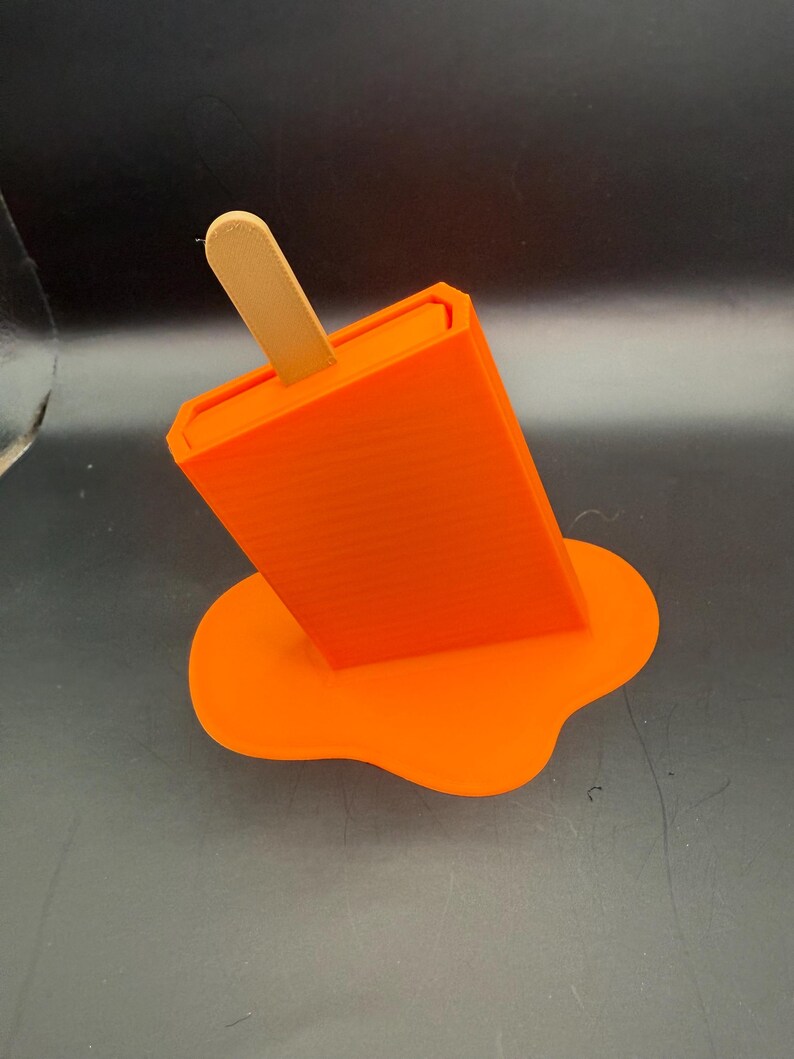 Peut inclure: Un support de t&eacute;l&eacute;phone en forme de glace &agrave; l'eau orange avec un b&acirc;ton en bois. Le support est con&ccedil;u pour ressembler &agrave; une glace &agrave; l'eau en train de fondre, avec la &laquo; cr&egrave;me glac&eacute;e &raquo; orange qui d&eacute;gouline sur la surface. Le support est en plastique.