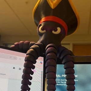 Puede incluir: Una caprichosa figura de pulpo morado oscuro con un sombrero de pirata con borde amarillo y una cinta roja para la cabeza. El pulpo está posado encima de un monitor de ordenador, con sus tentáculos colgando. El pulpo tiene una expresión severa.