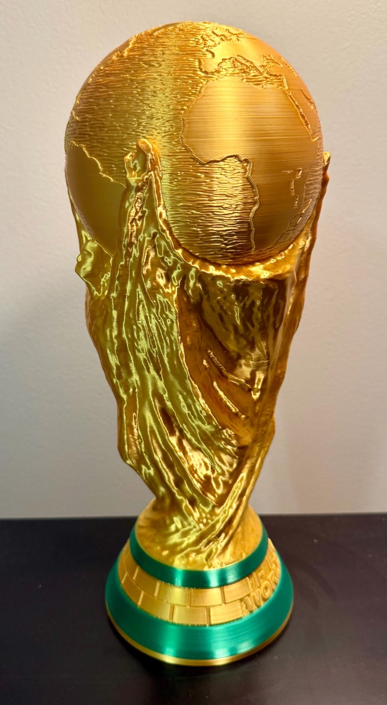May include: A gold-coloured trophy featuring a globe at the top supported by textured, flowing vertical elements. The base is circular with a green band and a gold-coloured section featuring the embossed text "FIFA WORLD CUP" in capital letters.