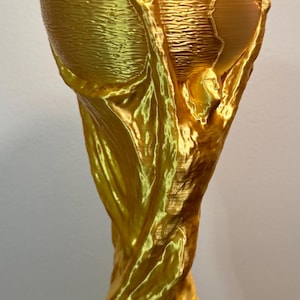 May include: A gold-coloured trophy featuring a stylised figure with arms raised, supporting a globe at the top. The base is circular with a green band and a brick-like pattern. The entire object has a visible 3D-printed texture throughout.
