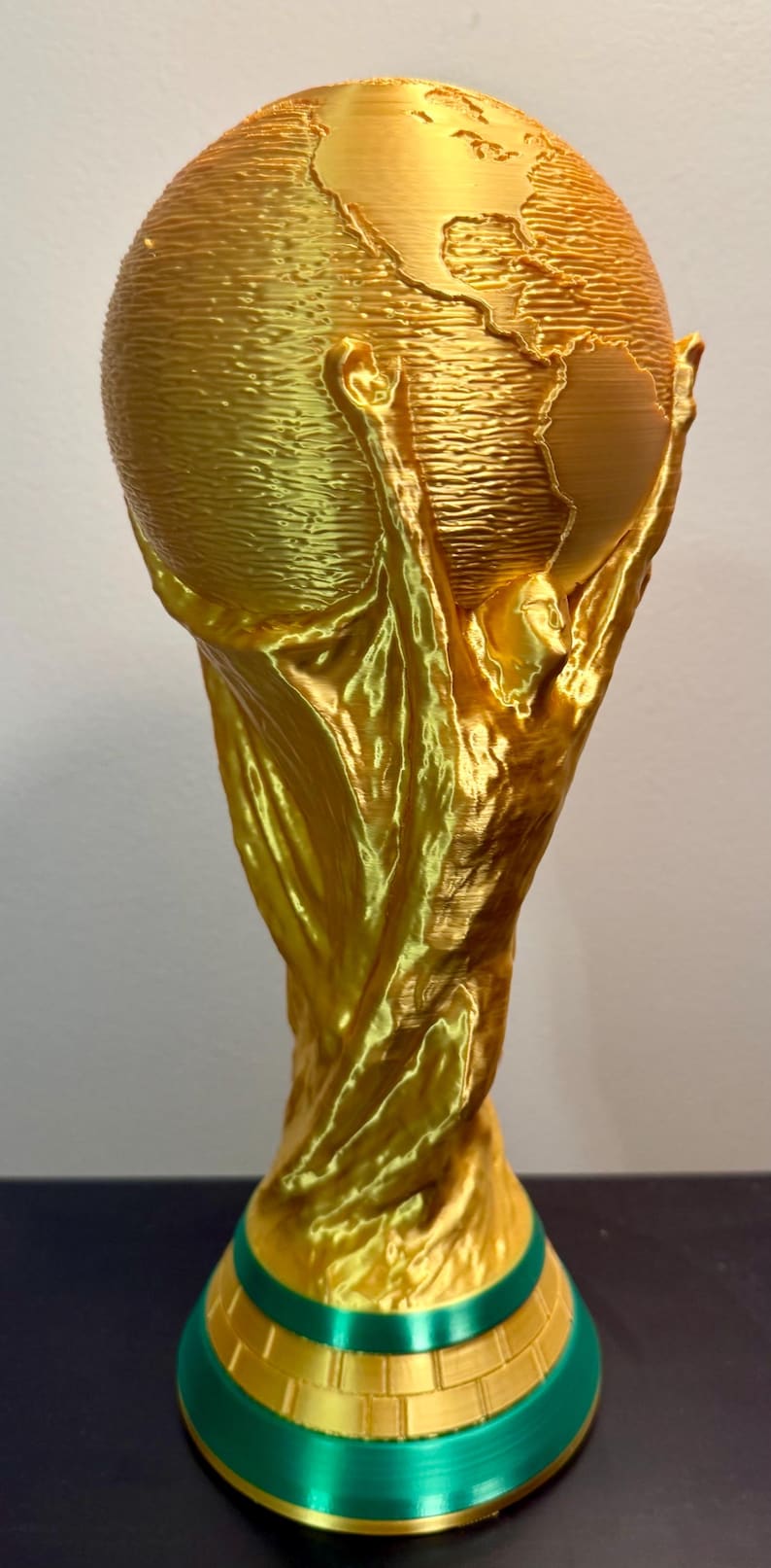 May include: A gold-coloured trophy featuring a stylised figure with arms raised, supporting a globe at the top. The base is circular with a green band and a brick-like pattern. The entire object has a visible 3D-printed texture throughout.