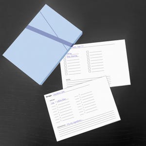 Könnte beinhalten: Eine hellblaue rechteckige Schachtel mit einem diagonalen Band, begleitet von zwei weißen Papierkarten. Eine Karte zeigt eine Checkliste mit dem Text "Deep cleaning" und "Home, Sept 01". Die andere ist eine Rezeptvorlage mit dem Text "Recipe: Chocolate Cake", "Amount", "White flour" und "Preparation: Mix dry ingredients".