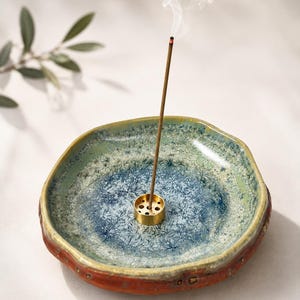 May include: A round, shallow ceramic incense burner with a crystalline glaze in shades of blue and green. The exterior has a textured, reddish-brown finish. A small, cylindrical brass incense holder sits in the center, holding a lit incense stick.