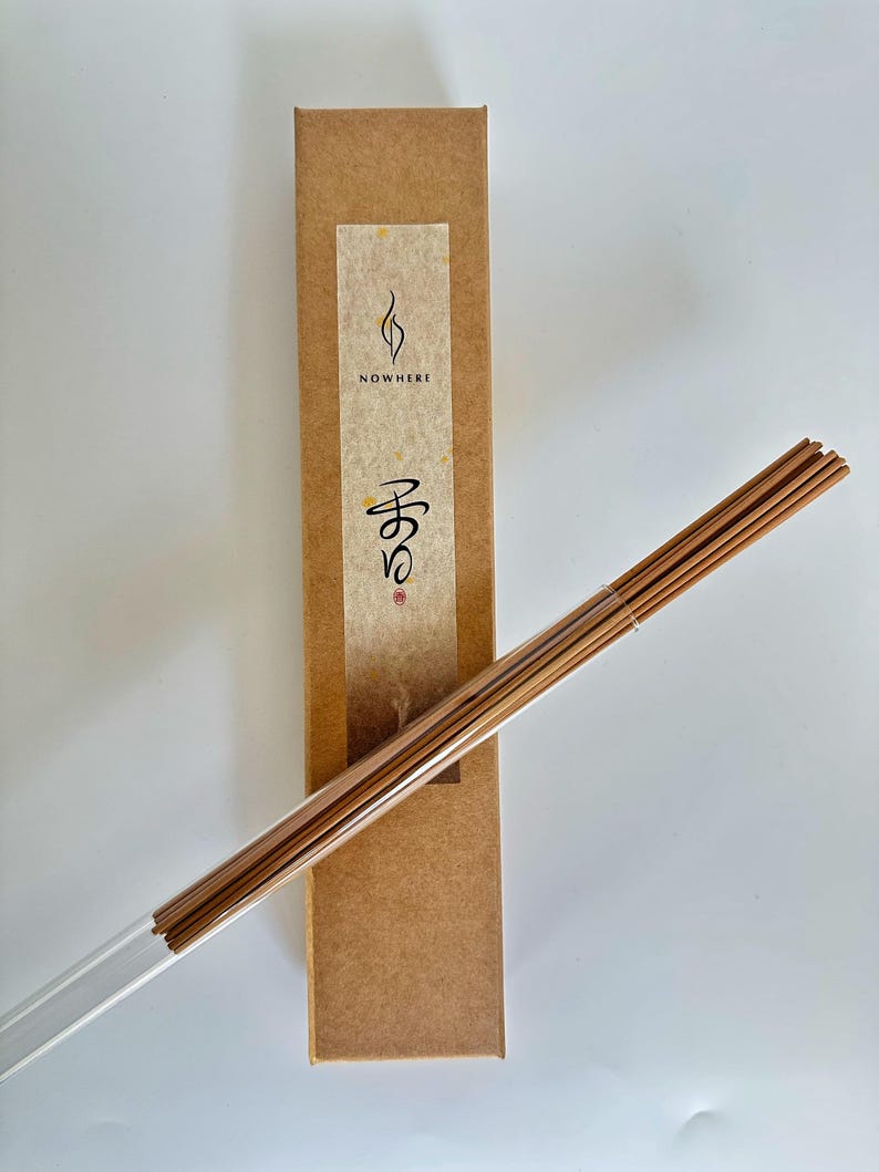 May include: A rectangular brown box with the word "NOWHERE" and Japanese text on a cream-colored label. A bundle of incense sticks is in a clear plastic tube, angled across the image.