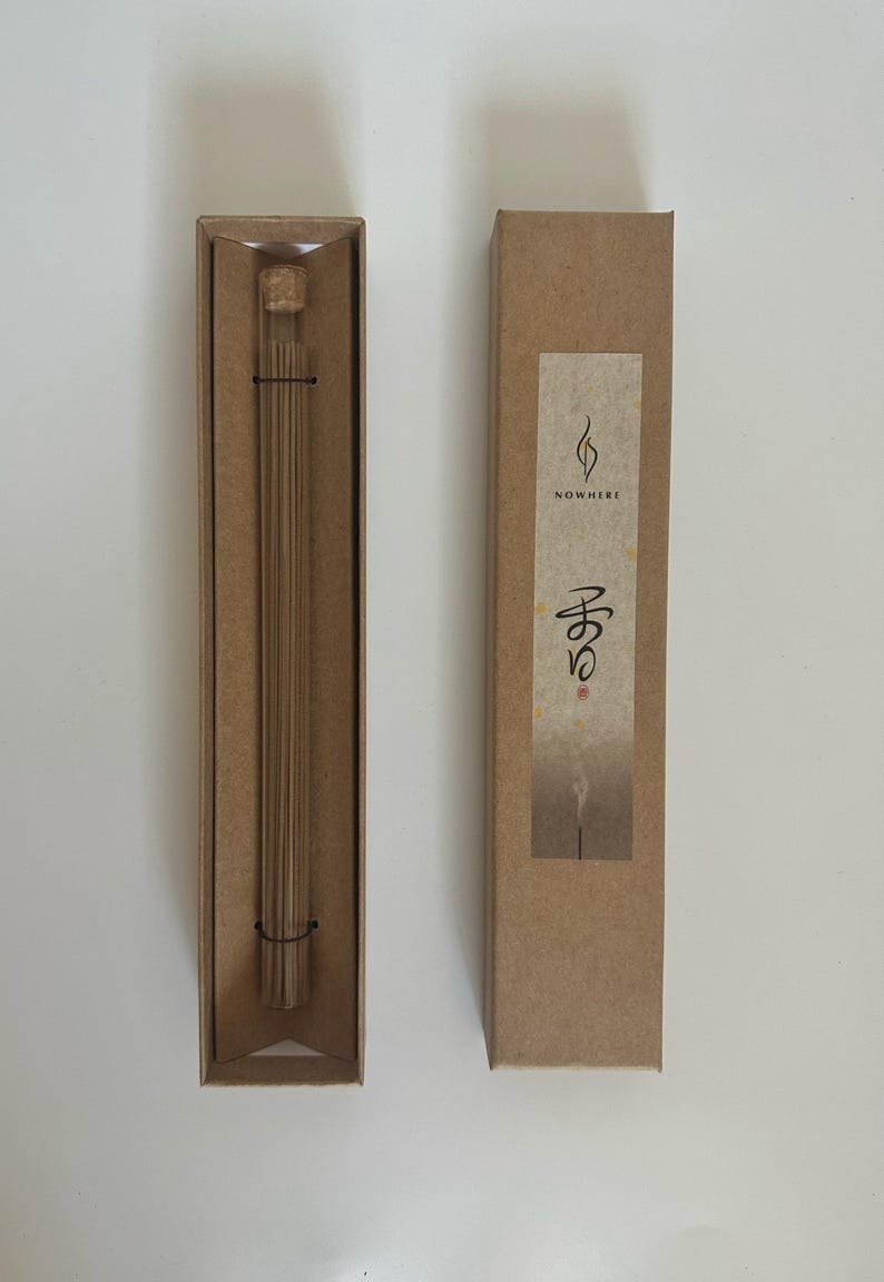 May include: Two rectangular brown cardboard boxes, one open and one closed, on a white surface. The open box contains a glass tube with incense sticks. The closed box has a label with the word "NOWHERE" and Japanese characters.