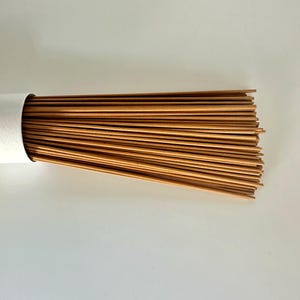 May include: A bundle of brown incense sticks emerging from a white cylindrical container. The incense sticks are straight and parallel, creating a textured, linear pattern. The background is a plain white surface.