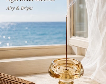 May include: A bundle of thin brown incense sticks next to a translucent amber glass incense holder with a single burning stick emitting a thin trail of white smoke. The text reads Premium Nha Trang Agarwood Incense and Airy & Bright.
