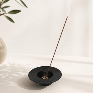 May include: A black, circular incense holder with a ridged design. A lit incense stick emits a thin stream of smoke. The holder sits on a white surface, with a vase and leafy branch in the background.