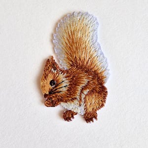 May include: Embroidered squirrel patch in brown, tan, and white. The patch shows a side profile of a squirrel with a bushy tail and detailed stitching. Suitable for application on clothing or accessories.