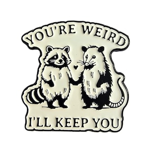 May include: A cream-coloured enamel pin featuring a raccoon and an opossum holding hands. The pin has black outlines and text that reads "YOU'RE WEIRD I'LL KEEP YOU". The design includes a small heart between the animals.