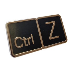 May include: A black and gold enamel pin shaped like the Ctrl and Z keys from a keyboard. The pin has a gold border and the letters Ctrl and Z are in gold.