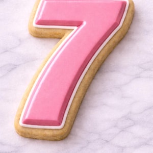 Peut inclure: Un biscuit en forme de chiffre sept, avec un glaçage rose, une bordure blanche et beige. Le biscuit est posé sur un fond en marbre blanc. Le biscuit est de couleur beige clair.