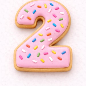 Puede incluir: Una galleta con forma de número dos, con glaseado rosa y chispas de colores. La galleta tiene un borde marrón claro. Las chispas son de varios colores, incluyendo azul, amarillo, verde y naranja.