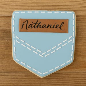 Puede incluir: Una galleta azul claro con forma de bolsillo de mezclilla, con detalles de costura blancos. Un parche rectangular marrón en la parte superior muestra el nombre "Nathaniel" en escritura cursiva negra. La galleta está sobre un fondo de madera.