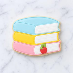以下が含まれることがあります： ロイヤルアイシングで装飾された3冊の本が積み重なった形のクッキー。一番上の本は水色、真ん中は黄色、一番下はピンク色です。各本には白いページがあり、一番下の本には赤いリンゴが付いた小さな緑色のしおりが付いています。