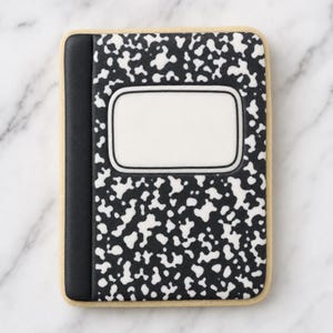 May include: A rectangular biscuit decorated with icing to resemble a composition notebook. The design features a black spine and a black and white speckled pattern across the cover, with a blank white rectangular label centred on the front.