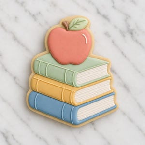 May include: A sugar biscuit shaped like a stack of three books with a red apple on top. The books are coloured light green, yellow, and blue, each featuring white icing details for the pages and spine. The apple has a small light green leaf.