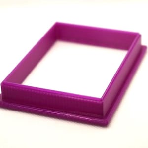 May include: A purple plastic rectangle cookie cutter with a smooth, raised edge.