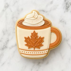 May include: A sugar cookie shaped like a mug filled with a beverage topped with white whipped cream. The mug is cream-colored with a brown handle and rim, featuring a prominent brown maple leaf design on the front and a dotted pattern at the base.
