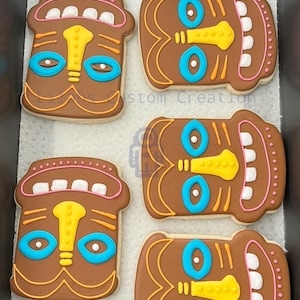 Puede incluir: Cuatro galletas de azúcar moreno decoradas con glaseado de colores para parecerse a máscaras tiki. Las máscaras tienen ojos azules, narices amarillas y dientes blancos.