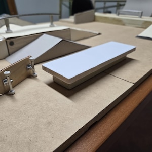 May include: A miniature skateboard with a natural wood deck and silver trucks sits near a white-topped platform. The scene includes other small ramps and rails, suggesting a fingerboard skate park setup. The background is a light brown surface.