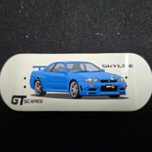 Könnte beinhalten: Ein weißes Fingerboard mit einem blauen Sportwagen-Design. Das Wort "SKYLINE" ist über dem Auto aufgedruckt, und "GT SCAPES" darunter. Das Board hat zwei kleine Löcher.