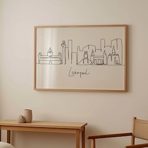 May include: A framed black and white line drawing of the Liverpool skyline. The drawing is on a white background and the frame is a light wood color. The drawing is hanging on a white wall.