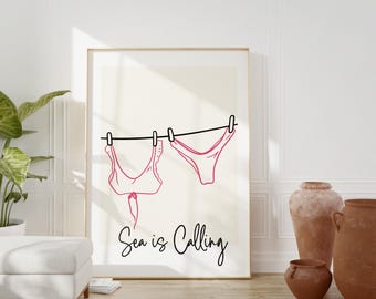 May include: A white framed print with a black line drawing of pink bikini tops and bottoms hanging on a clothesline. The text "Sea is Calling" is written below the image.