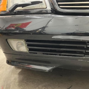 Puede incluir: Un coche negro con un parachoques delantero que tiene una pequeña luz antiniebla rectangular transparente en el lado izquierdo. El parachoques tiene una rejilla de plástico negro con lamas horizontales.