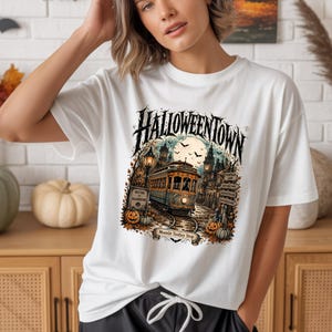 May include: A white short-sleeve t-shirt featuring a graphic print of a trolley in a town named Halloweentown. The design includes a full moon, bats, pumpkins, and street signs. The text reads HALLOWEENTOWN and Moonlit Trolley Stop.