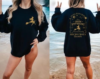 May include: Black sweatshirt with a gold witch on a broom design on the front. The back of the sweatshirt features text that reads "The Salem Apothecary Massachusetts" with additional text and a witch graphic. The sweatshirt is long-sleeved.