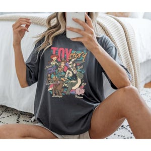 A person is sitting on a bed, wearing a black t-shirt with a colorful graphic of characters from the Toy Story movie.