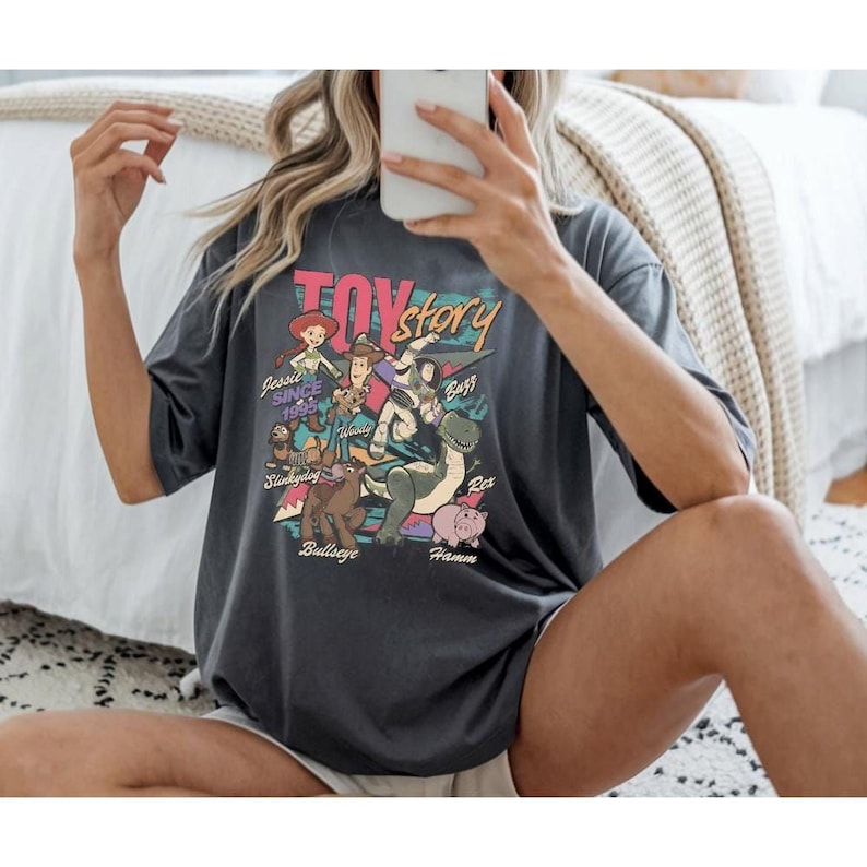 A person is sitting on a bed, wearing a black t-shirt with a colorful graphic of characters from the Toy Story movie.