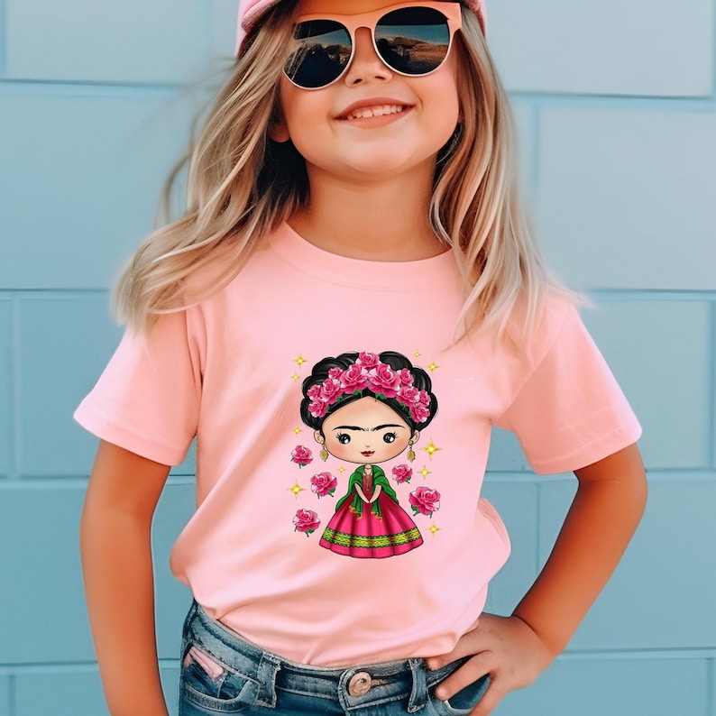 Puede incluir: Una camiseta rosa claro con una ilustraci&oacute;n de dibujos animados de una mujer con una corona floral y un vestido rosa. El dise&ntilde;o est&aacute; rodeado de rosas rosas y estrellas doradas. La ni&ntilde;a lleva gafas de sol y una gorra rosa.